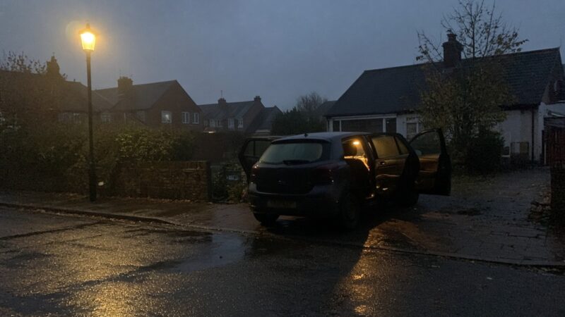 Blue hour suburban street after rain with a dark hatchback parked doors open and warm interior light on wet tarmac under an orange streetlamp