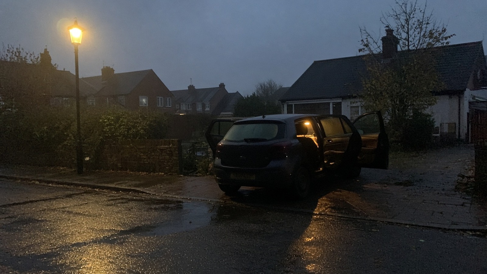 Blue hour suburban street after rain with a dark hatchback parked doors open and warm interior light on wet tarmac under an orange streetlamp